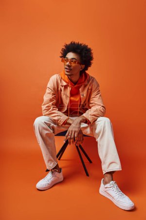 Ein trendiger junger afroamerikanischer Mann in stylischer Kleidung und Sonnenbrille sitzt auf einem Stuhl
