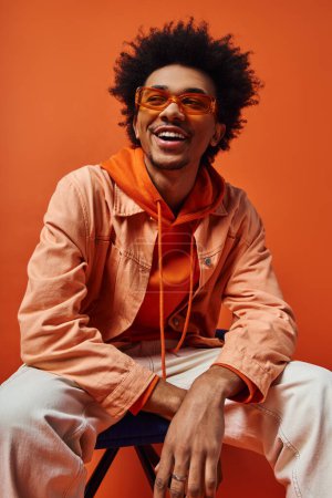 Téléchargez les photos : Jeune homme afro-américain aux cheveux bouclés assis en toute confiance sur une chaise, portant des lunettes de soleil élégantes sur fond orange. - en image libre de droit