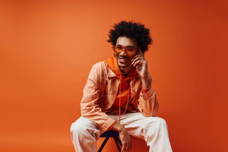 Foto de Un hombre afroamericano de moda con cabello rizado, gafas de sol y atuendo elegante se sienta en una silla sobre un fondo naranja. - Imagen libre de derechos
