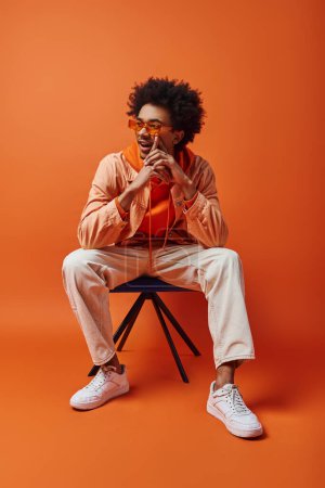Ein stilvoller junger afroamerikanischer Mann mit lockigem Haar und Sonnenbrille sitzt kühn auf einem Stuhl vor orangefarbenem Hintergrund..
