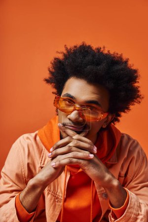 Ein trendiger junger afroamerikanischer Mann mit lockigem Haar, Brille und orangefarbener Jacke auf leuchtend orangefarbenem Hintergrund.