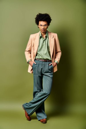 Foto de Un hombre afroamericano joven y elegante con el pelo rizado se para con confianza frente a una pared verde vibrante, con atuendo de moda y gafas de sol. - Imagen libre de derechos