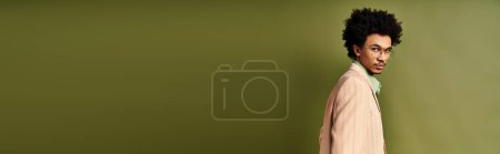 Téléchargez les photos : Un jeune homme afro-américain élégant avec des cheveux bouclés et une tenue tendance debout devant un mur vert vibrant. - en image libre de droit