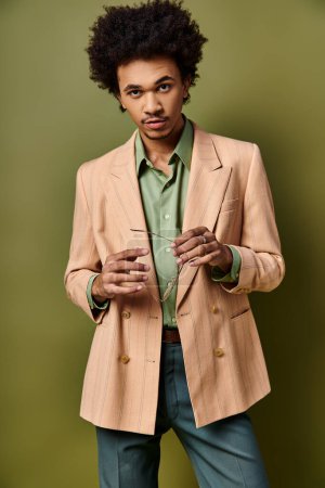 Jeune homme afro-américain élégant aux cheveux bouclés, vêtu d'une veste bronzée et d'une chemise verte, en tenue tendance et lunettes de soleil.