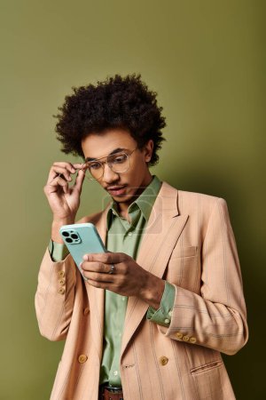 Foto de Un joven afroamericano con estilo en traje y gafas de sol, absorbido en su teléfono celular sobre un fondo verde. - Imagen libre de derechos