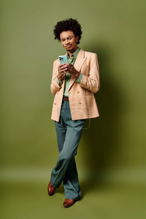 Ein stilvoller junger afroamerikanischer Mann im Anzug hält ein Handy vor grünem Hintergrund und strahlt Zuversicht und Raffinesse aus.