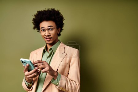 Foto de Un joven afroamericano con estilo en un traje y gafas de sol, absorbido en su teléfono celular sobre un fondo verde. - Imagen libre de derechos