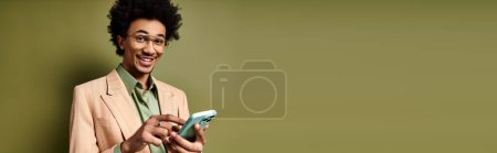 Foto de Elegante joven afroamericano con un traje y gafas de sol sosteniendo un teléfono celular sobre un fondo verde. - Imagen libre de derechos