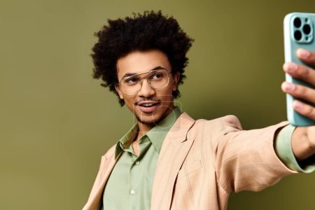 Téléchargez les photos : Un jeune homme afro-américain élégant avec des cheveux bouclés et des lunettes de soleil prenant un selfie avec son téléphone portable sur un fond vert. - en image libre de droit
