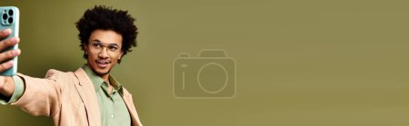 Photo for A young African American man, stylishly dressed, takes a selfie with his cellphone against a green background. - Royalty Free Image