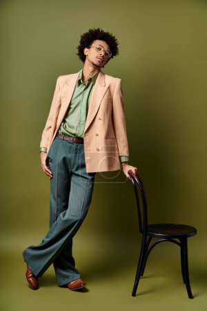 Ein trendiger, lockiger afroamerikanischer Mann in stylischer Kleidung steht neben einem Stuhl vor einer leuchtend grünen Wand.