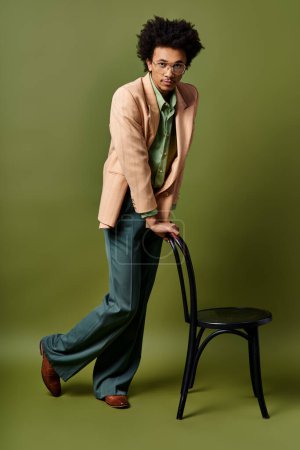 Foto de Un hombre afroamericano elegante y rizado se apoya pensativamente en una silla, usando atuendos de moda y gafas de sol contra un fondo verde.. - Imagen libre de derechos