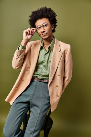 Téléchargez les photos : Un jeune homme afro-américain élégant en costume et lunettes de soleil, assis sur une chaise sur un fond vert. - en image libre de droit