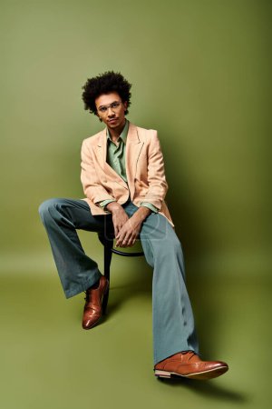 Photo for Stylish young African American man with curly hair, sunglasses, and trendy attire sitting on a chair with his legs crossed. - Royalty Free Image