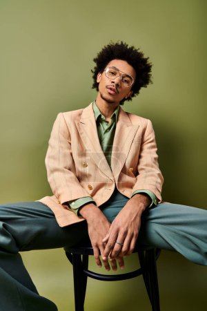 A stylish young African American man with curly hair sitting on a chair, legs crossed, in trendy attire and sunglasses on a green background.