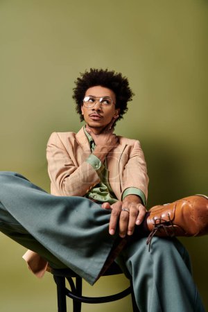 A stylish young African American man with curly hair sits confidently atop a wooden chair against a green background.