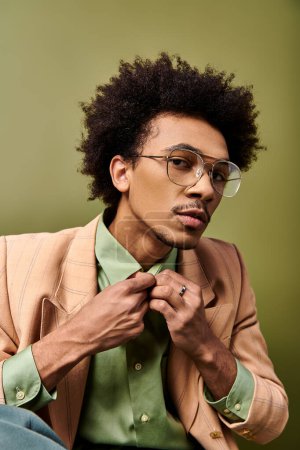 Un jeune homme afro-américain élégant en costume et lunettes boutonne méticuleusement sa chemise sur un fond vert vif.
