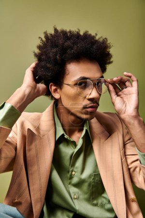 Stylish young African American man with curly hair, sharp suit, and sunglasses, sitting on a green background.
