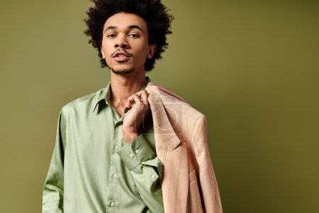 Stylish young African American man with curly hair holds a jacket over his shoulder, wearing trendy attire on a green background.