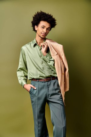 A stylish young African American man with curly hair, wearing a green shirt and blue pants, poses confidently against a green background.