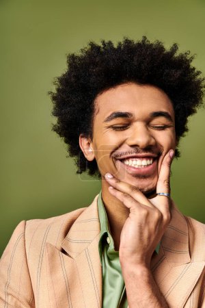 Téléchargez les photos : Jeune homme afro-américain élégant aux cheveux bouclés et aux vêtements tendance souriant pour la caméra sur un fond vert vibrant. - en image libre de droit