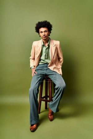 A trendily dressed young African American man with curly hair sitting atop a wooden stool against a green background.