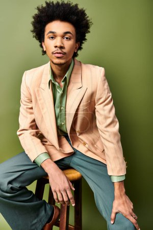 Téléchargez les photos : Jeune homme afro-américain élégant aux cheveux bouclés assis sur un tabouret en costume tendance sur un fond vert. - en image libre de droit