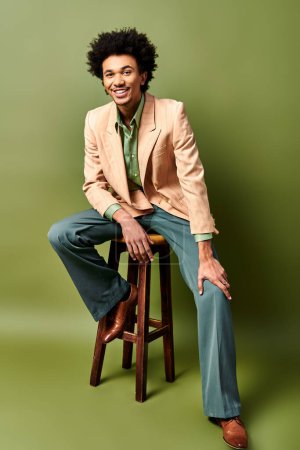 A stylish young African American man with curly hair sitting on top of a wooden stool against a green background.