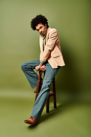 A stylish young African American man in trendy attire sitting on top of a wooden chair against a green background.