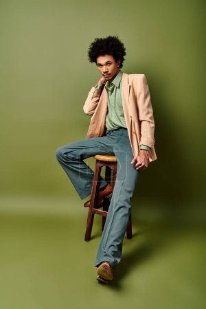 A young, curly African American man in stylish attire sits gracefully atop a wooden stool against a vibrant green background.