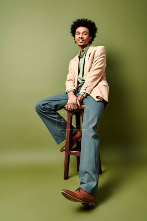 A stylish young African American man with curly hair sits confidently on a wooden stool, wearing trendy attire