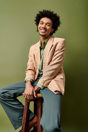 A stylish young African American man with curly hair sits atop a wooden stool, donning trendy attire on a green background.
