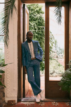 Schöner afroamerikanischer Mann im blauen Anzug, der in einer Tür steht, die von üppigem Grün umgeben ist.