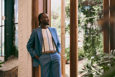 A handsome African American man in a blue suit leaning against a wall in a lush green garden.