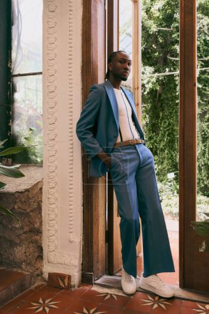Téléchargez les photos : Beau Afro-Américain en costume bleu appuyé contre une porte de jardin vert vif. - en image libre de droit