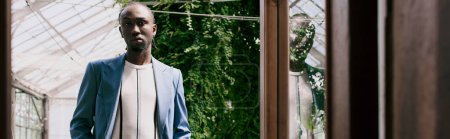 Foto de Un apuesto hombre afroamericano de sofisticado estilo elegante se encuentra frente a una ventana, enmarcada por un jardín verde vívido. - Imagen libre de derechos