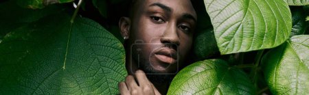 A sophisticated African American man hides behind a large green leaf in a vibrant garden.