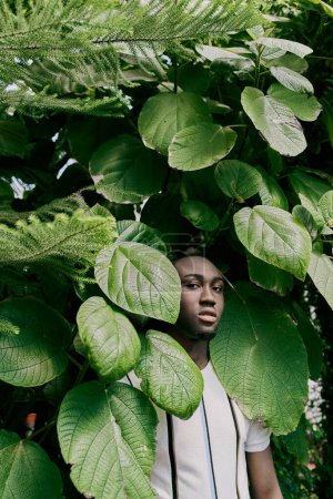 Foto de Un hombre afroamericano con estilo se para con confianza frente a un exuberante árbol verde. - Imagen libre de derechos