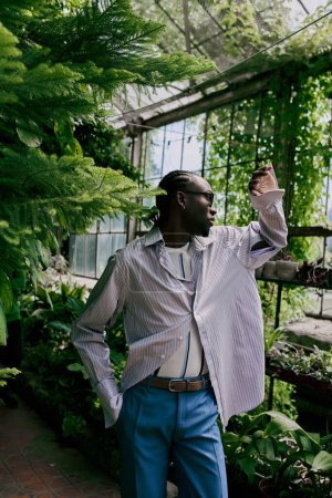 Handsome African American man in white shirt and blue pants poses in lush greenhouse.