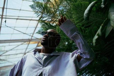 Foto de Un hombre afroamericano sofisticado posa frente a una exuberante palmera. - Imagen libre de derechos