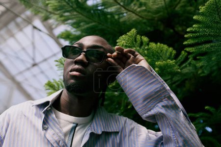 Photo for Handsome African American man in dapper style poses in a vivid green greenhouse. - Royalty Free Image
