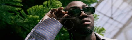 Téléchargez les photos : Un Afro-Américain en lunettes de soleil et tenue sophistiquée dans un jardin verdoyant animé. - en image libre de droit