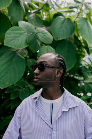 A handsome African American man with sophisticated style stands in front of a lush green tree, wearing sunglasses.