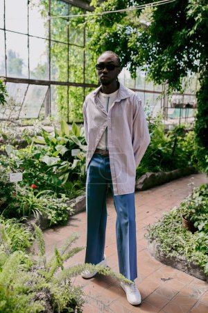 A stylish African American man stands confidently in front of a greenhouse filled with lush green plants.