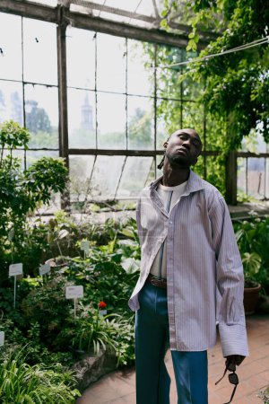 Handsome African American man in sophisticated attire standing in lush green greenhouse.