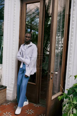 Handsome African American man in sophisticated dapper style stands in front of a glass door in a vivid green garden.