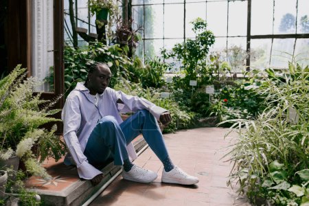 Handsome African American man in sophisticated dapper style sitting on a bench in a lush greenhouse.
