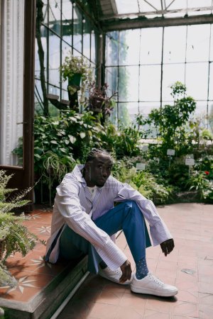 Handsome African American man in dapper style sitting on bench in vibrant greenhouse.