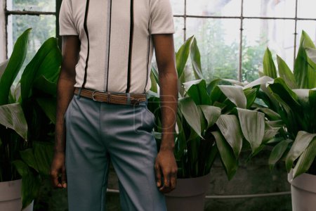 Téléchargez les photos : Un homme afro-américain élégant pose élégamment parmi une variété de plantes en pot dans un jardin luxuriant. - en image libre de droit