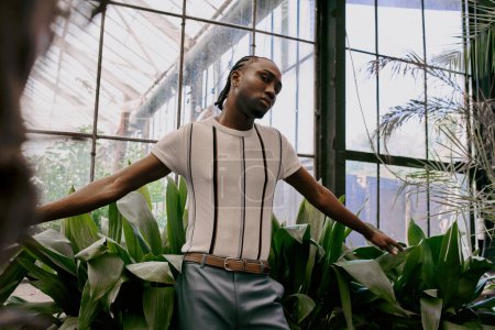 Téléchargez les photos : Bel homme afro-américain avec un style sophistiqué dapper debout devant une fenêtre dans un jardin vert vif. - en image libre de droit